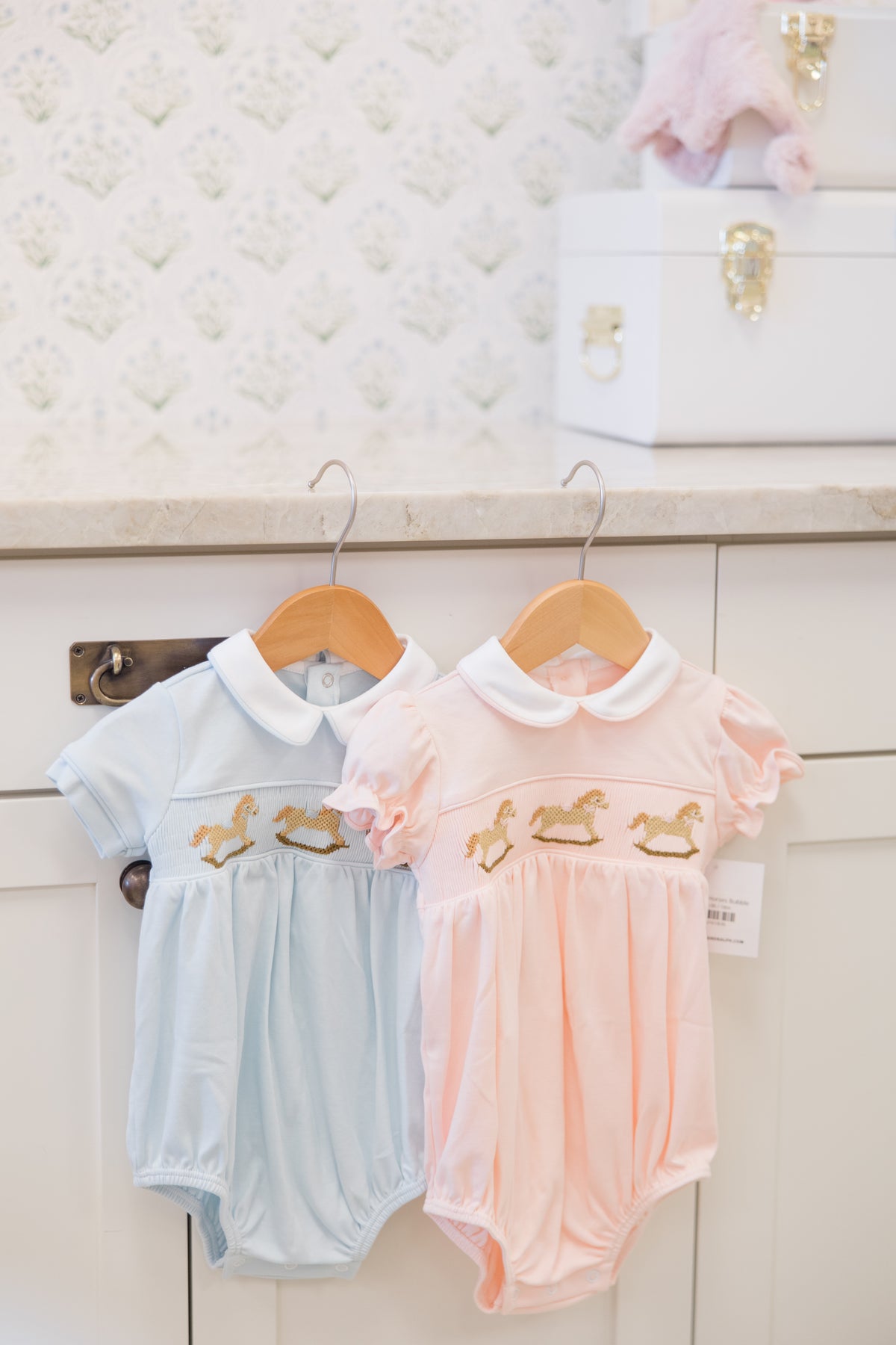 Two baby short-sleeve bodysuits hanging on wooden hangers against a white cabinet. One is light blue and the other is light pink, both with white Peter Pan collars and gold embroidered horses on the chest.