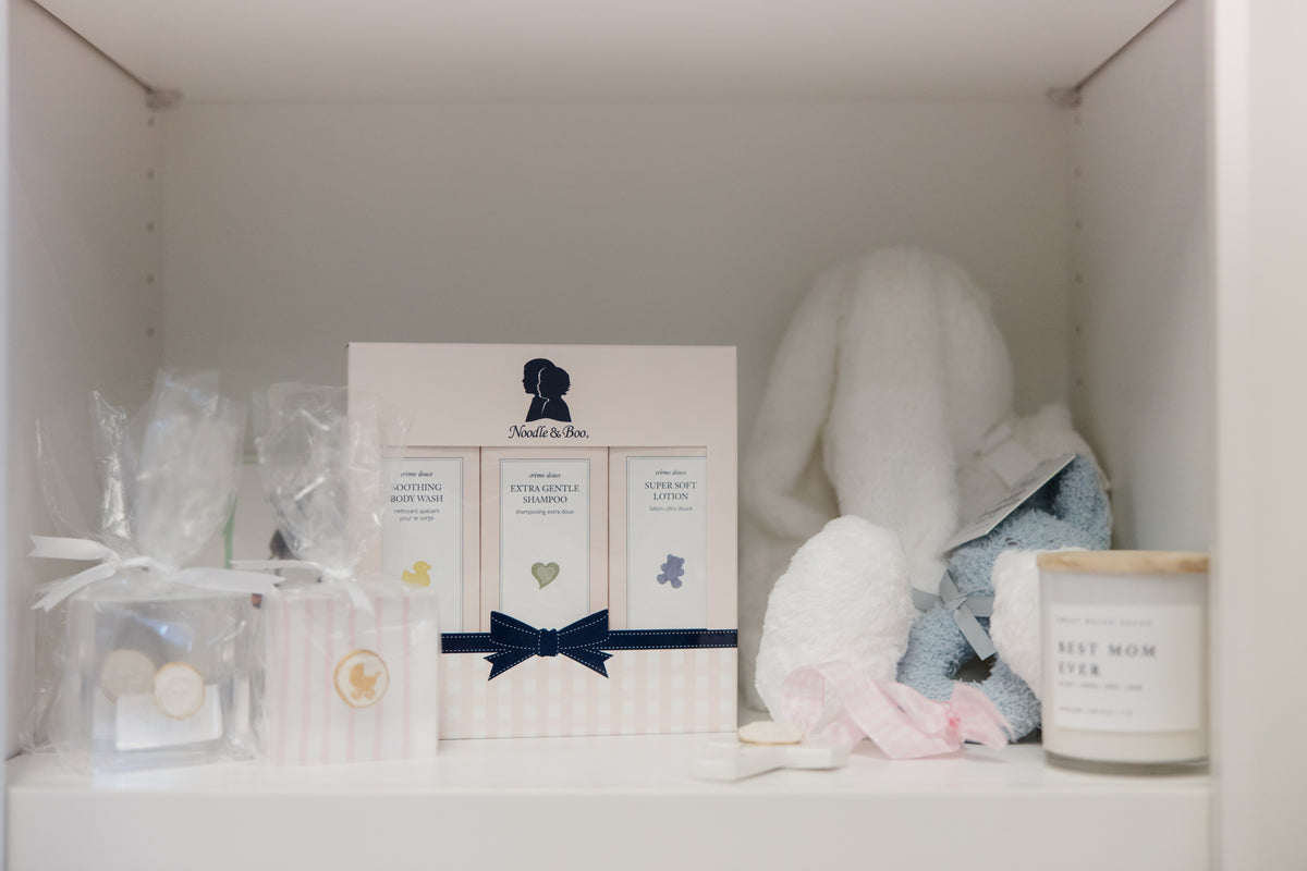 A baby shower gift set arranged on a white shelf, featuring a central white box with three panels and a navy ribbon, a plush white bunny, a blue and white stuffed toy, a candle, and small wrapped gift bags.